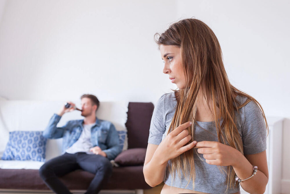 A concerned woman looking back at a man drinking on the couch.